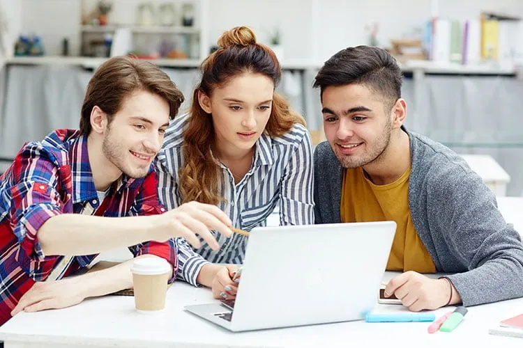 Three students working together on a laptop at a table with study materials and a coffee cup.